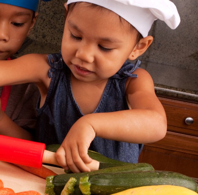 A toddler in a chef's hat carefully slicing zucchini with a child-safe knife while an older child steadies the board
