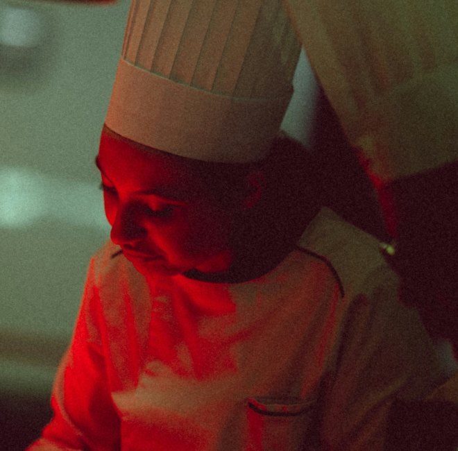 A chef in a white coat and tall toque working in a professional kitchen under warm red lighting