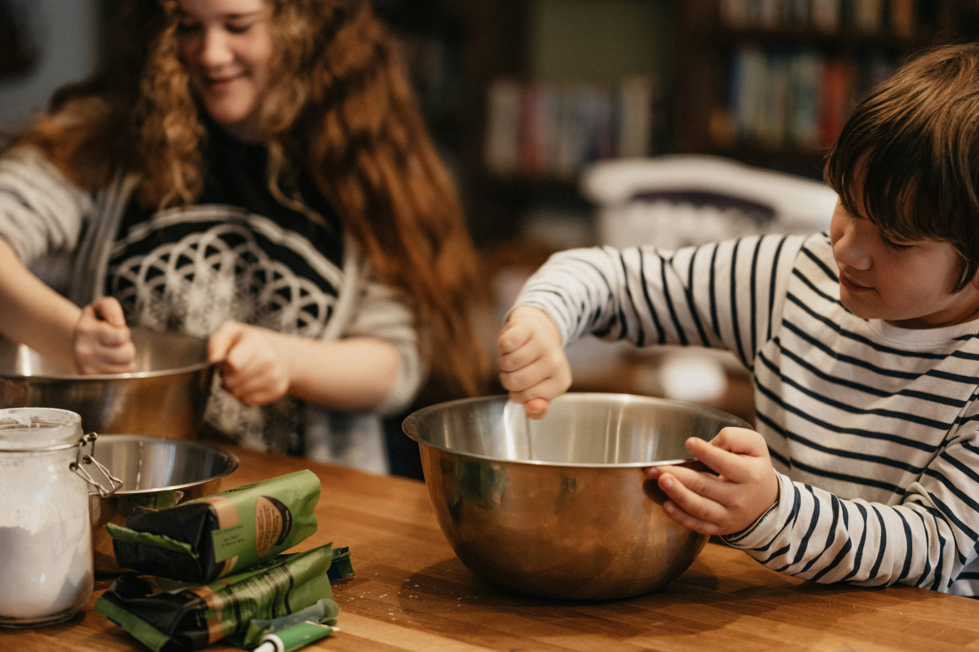 Two children side by side at a kitchen table, each stirring their own metal mixing bowl