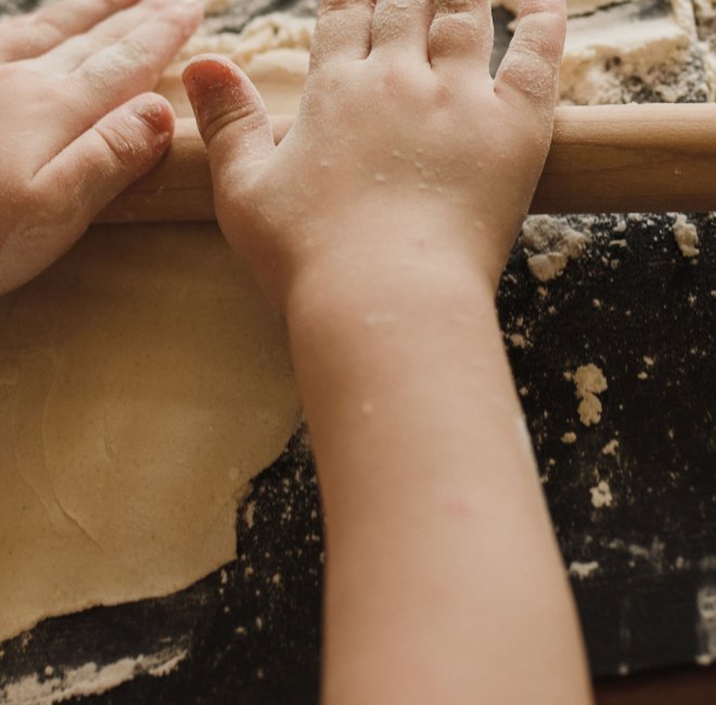 A child's flour-dusted hands rolling out dough with a wooden rolling pin, flour scattered across the counter