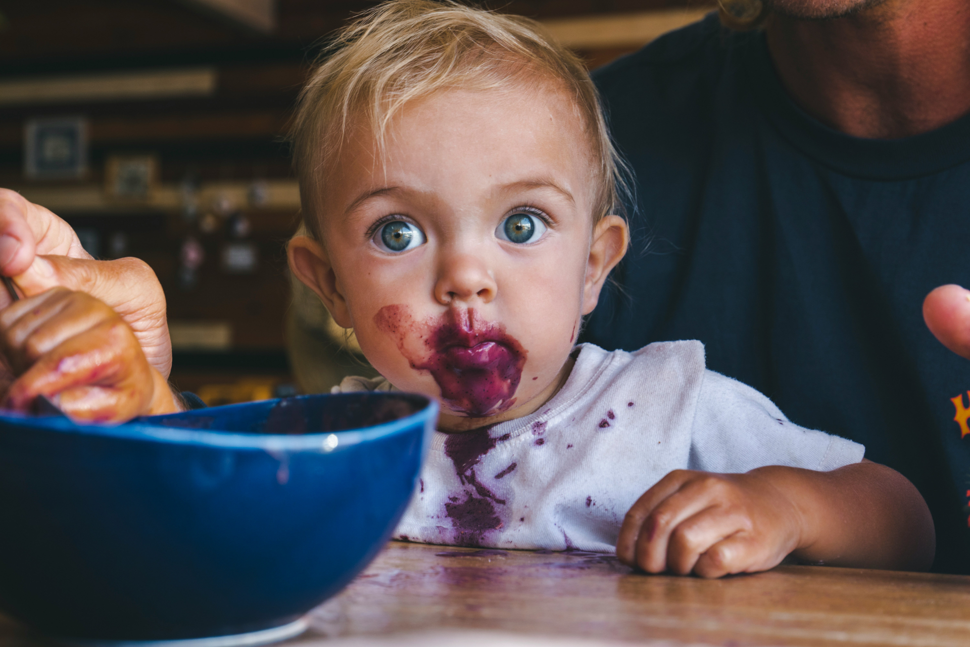 A wide-eyed blonde toddler with dark berry puree smeared across their face mid-meal, spoon and bowl in front of them